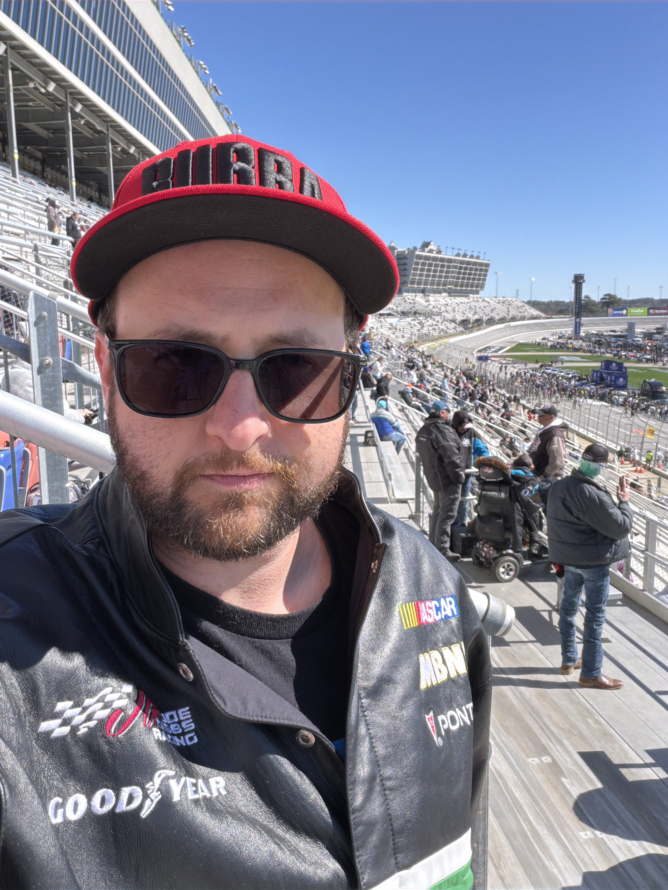 A thirtysomething man in a black leather Joe Gibbs Racing Bobby Labonte jacket and a red hat with “BUBBA” on it in black bubble letters, wearing transition glasses with the lenses fully blacked out. He is not smiling and taking a selfie in front of the grandstands at EchoPark Speedway in Atlanta.